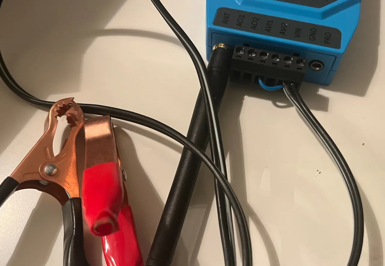 Ein blaues Funkmodul mit beschrifteten Klemmen und Antenne. Zwei Kabel mit roter und schwarzer Krokodilklemme sind am Spannungseingang angeschlossen.