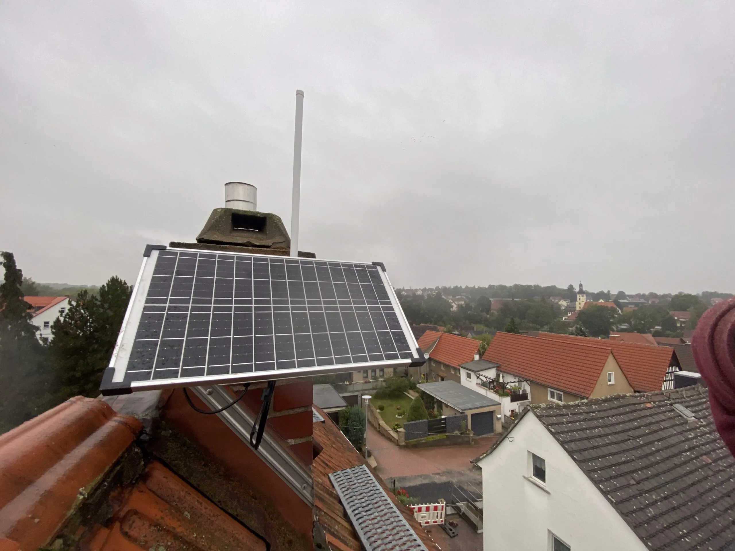 Ein kleines Solarmodul und ein Funkgerät auf einem Dachfirst, dahinter Dächer und ein Kirchturm.